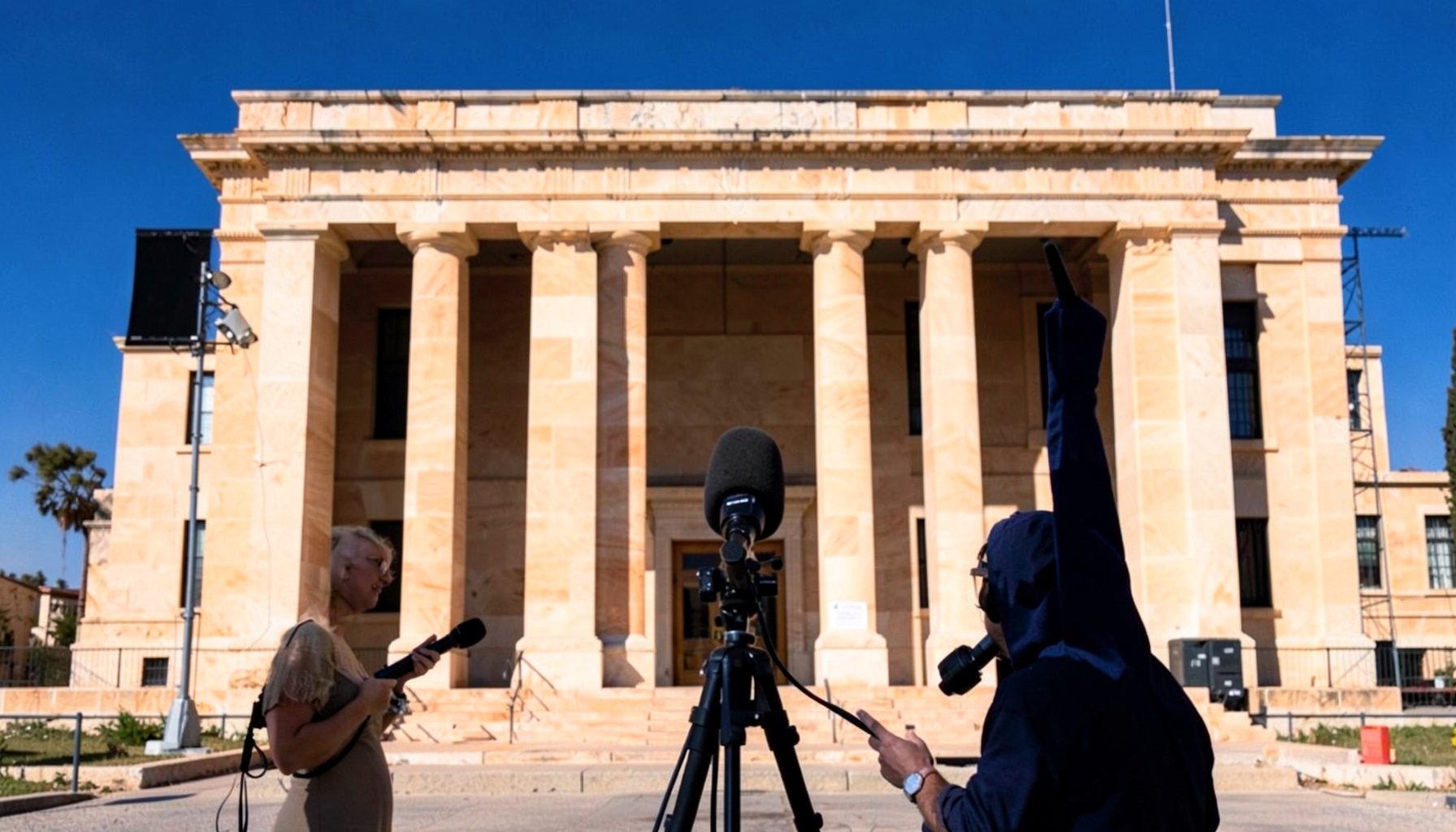 An outdoor broadcast setup with a reporter holding a microphone against a backdrop of a government building facade in warm sandstone tones under midday sun.