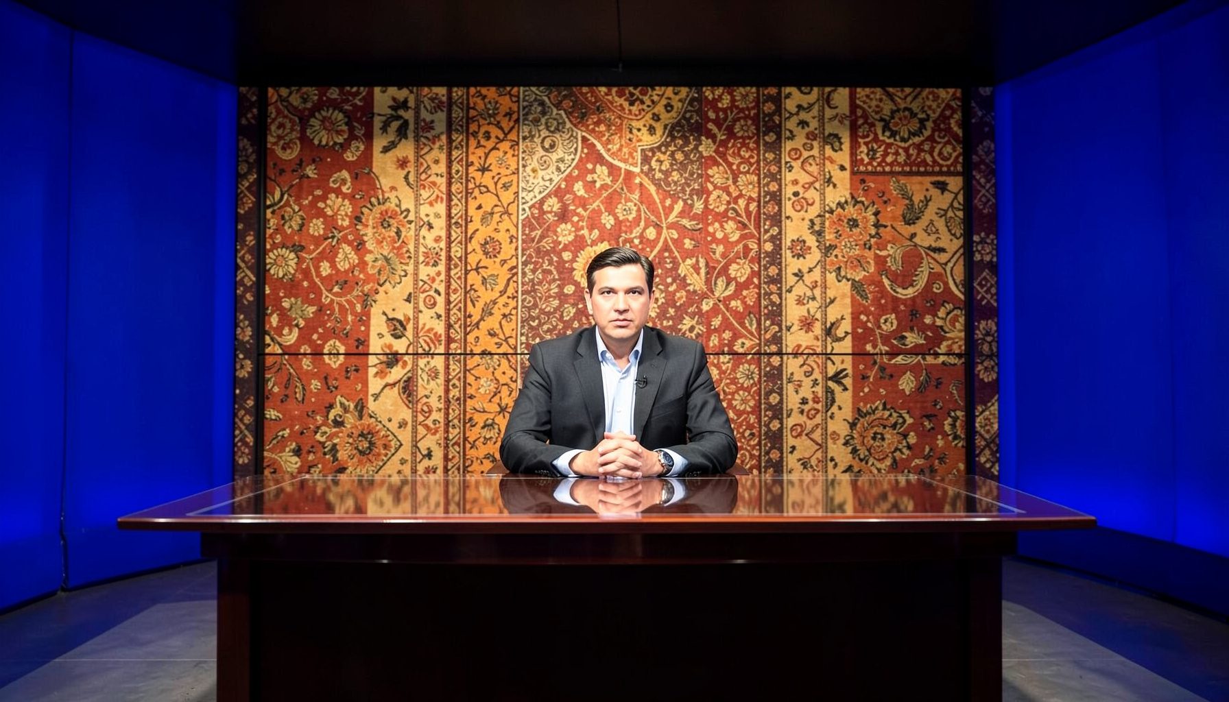 A regional news studio with traditional textile patterns on the backdrop in earthy orange and gold, and a presenter seated at a polished desk.