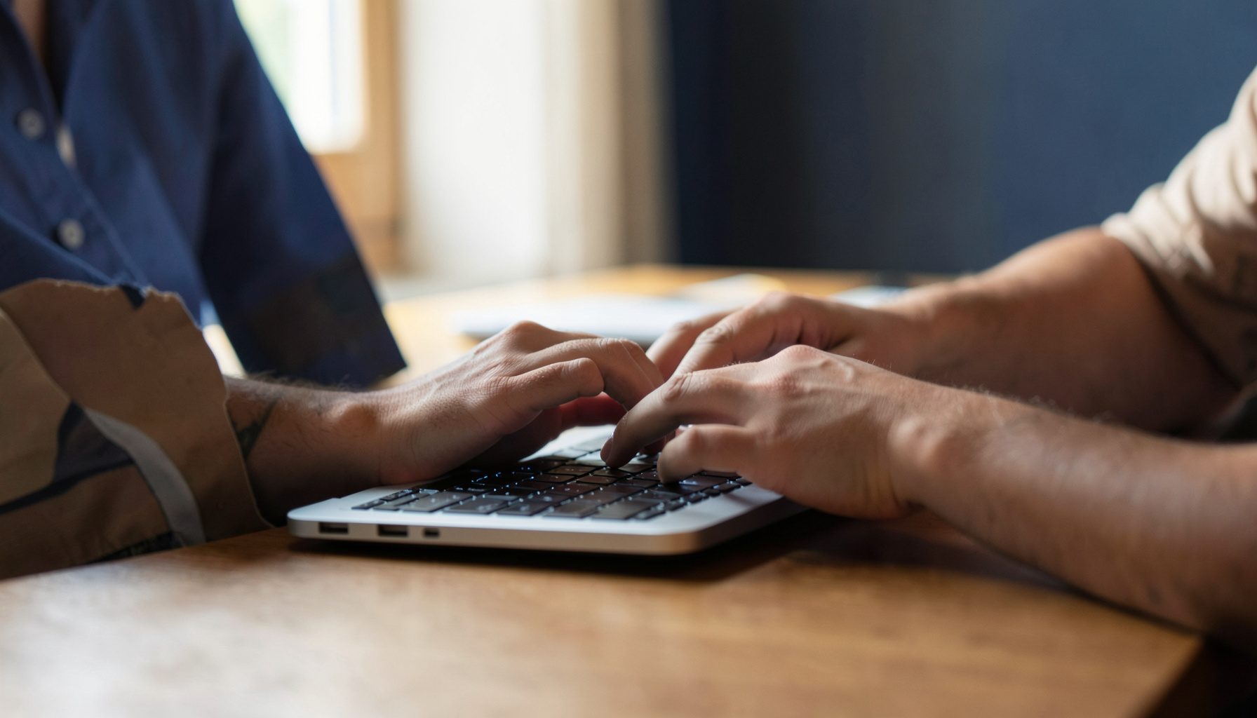 A close-up view of a person's hands working on a laptop at a wooden desk, with soft natural light from a window, warm beige and brown tones.
