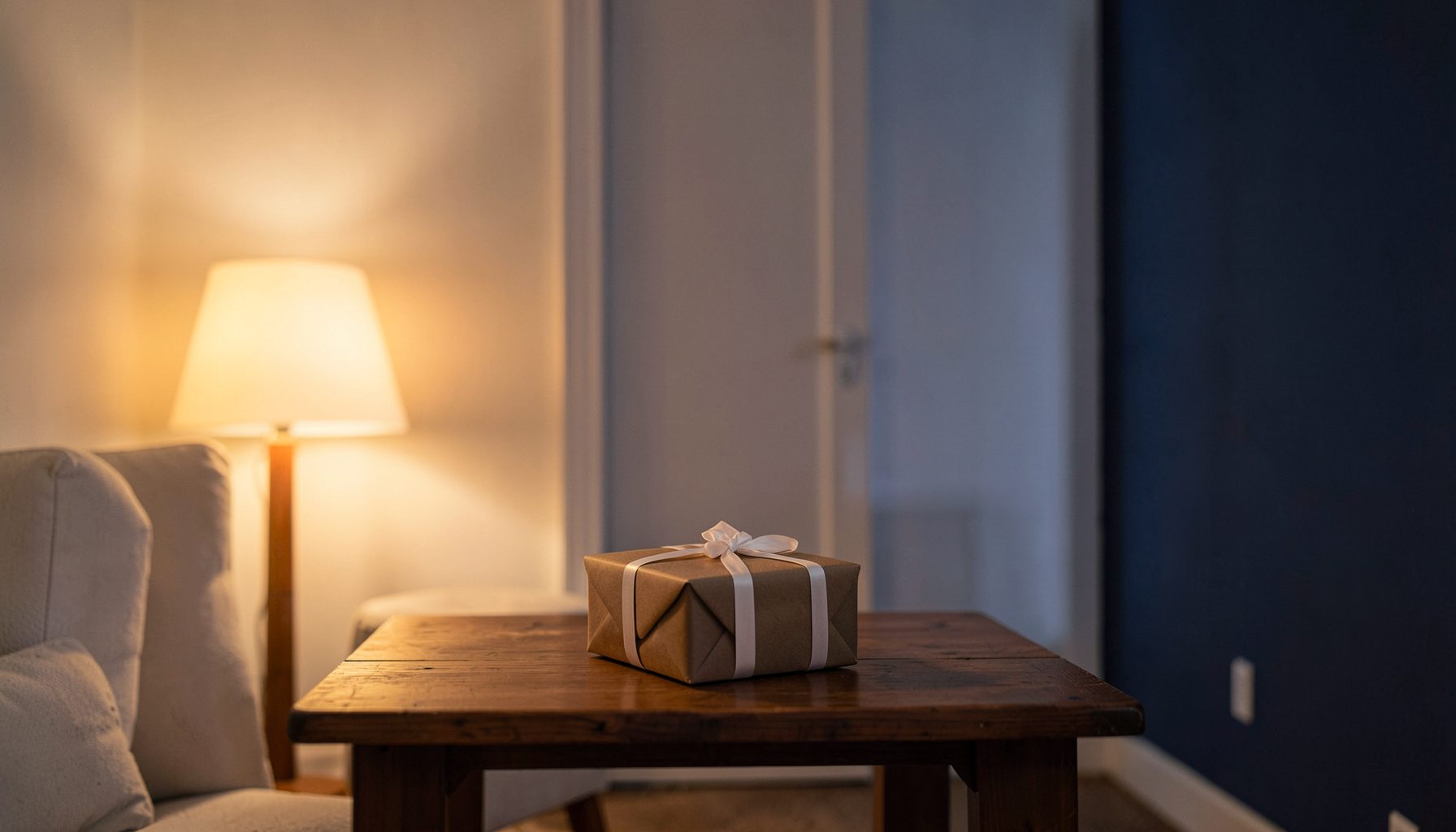 Cozy indoor setting with warm lamp light, a wrapped gift box on a wooden table, soft cream and brown tones