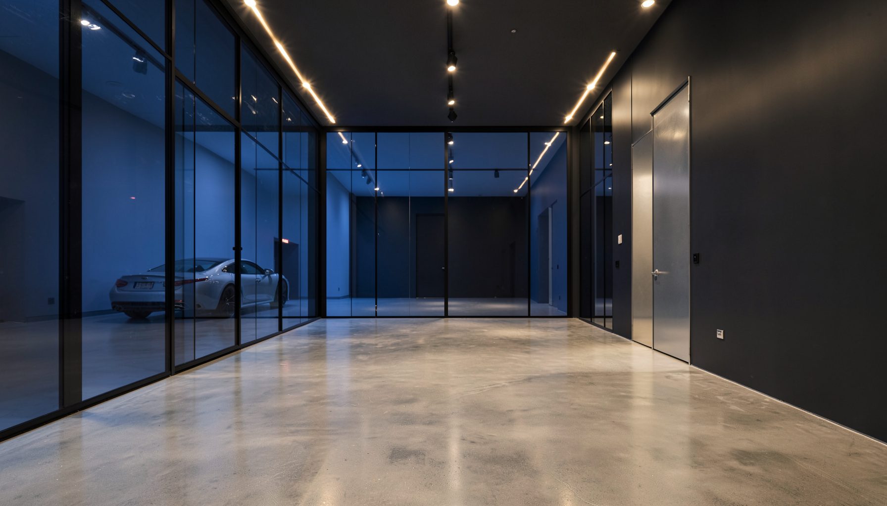 A sleek modern car showroom interior with polished concrete floors, floor-to-ceiling glass panels, and warm overhead lighting casting soft reflections across the space. The room is symmetrical and uncluttered, with deep charcoal and silver tones dominating.