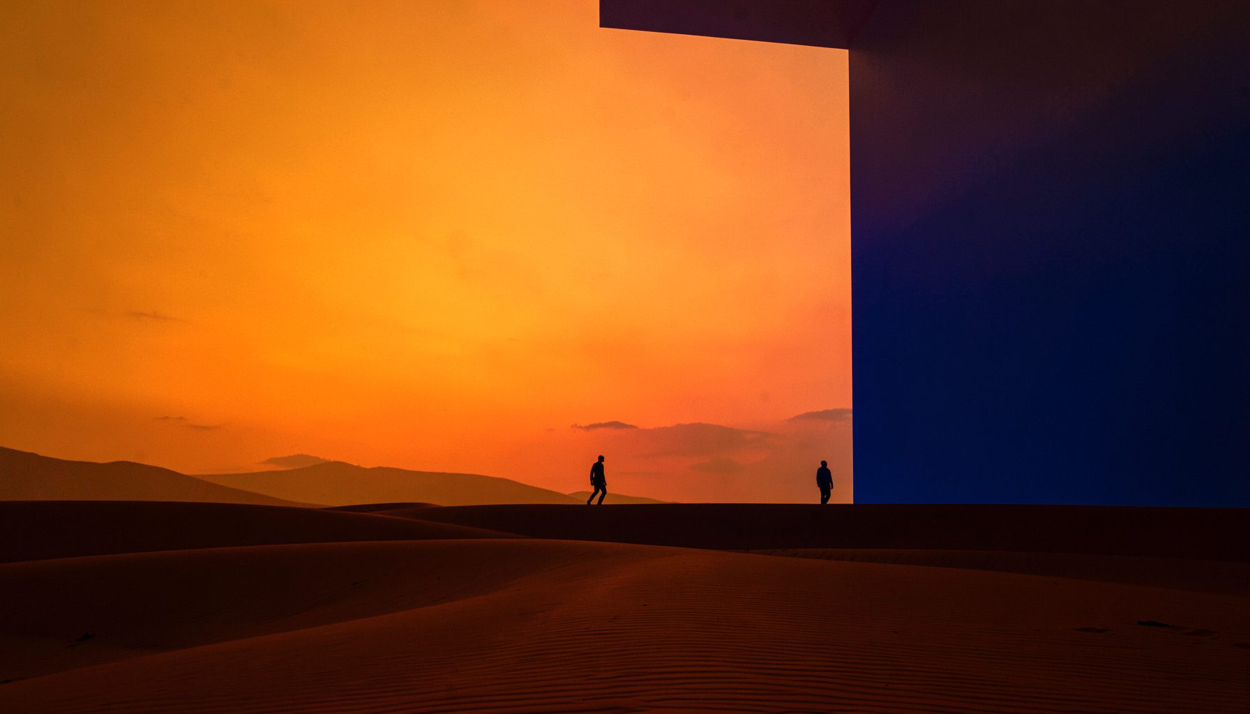 Dramatic wide shot of a desert landscape at golden hour with two figures silhouetted against a fiery orange sky, warm amber and sand tones