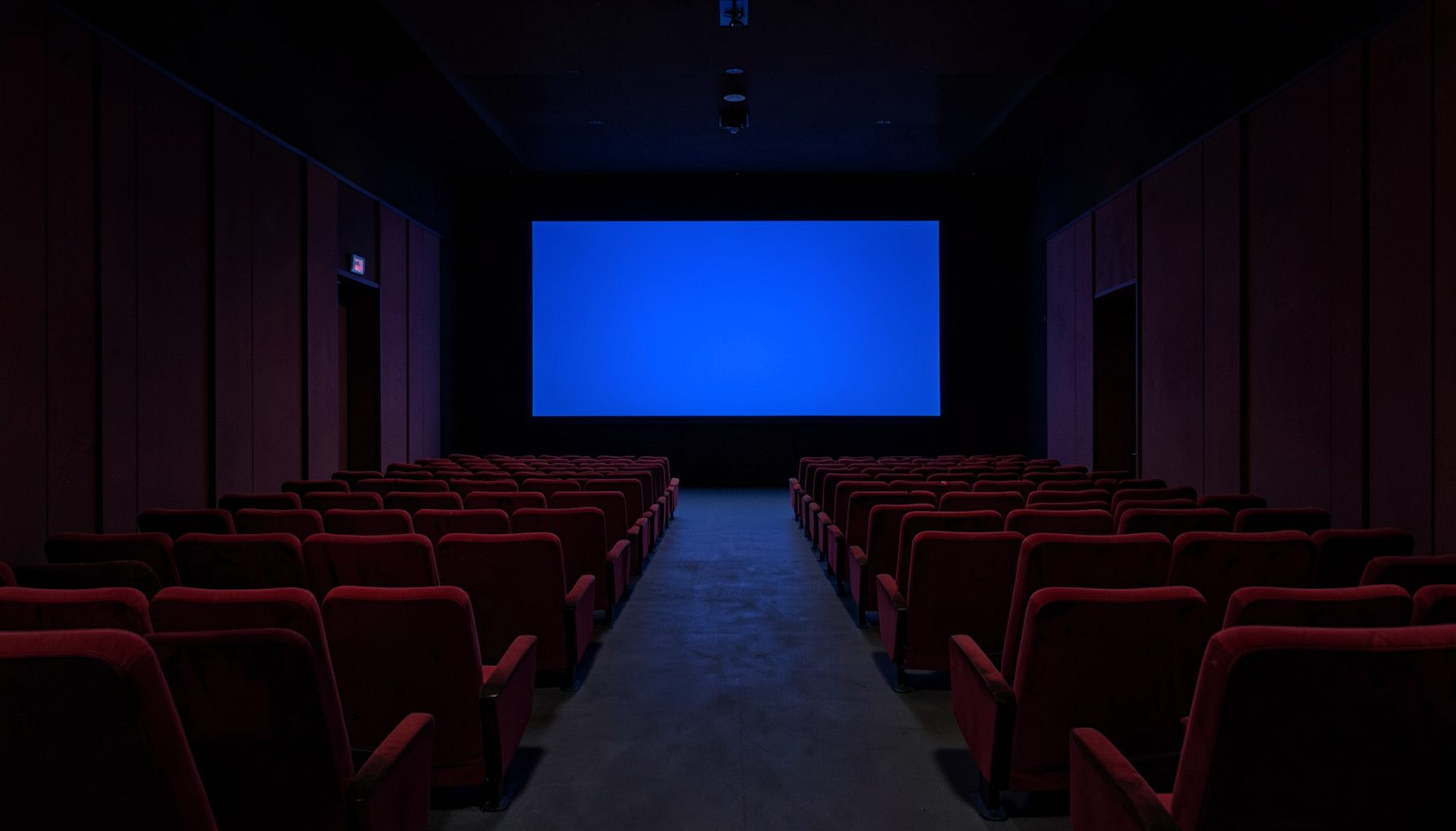 A dark, atmospheric cinema interior with rows of empty red velvet seats receding symmetrically toward a glowing screen, deep burgundy and charcoal tones