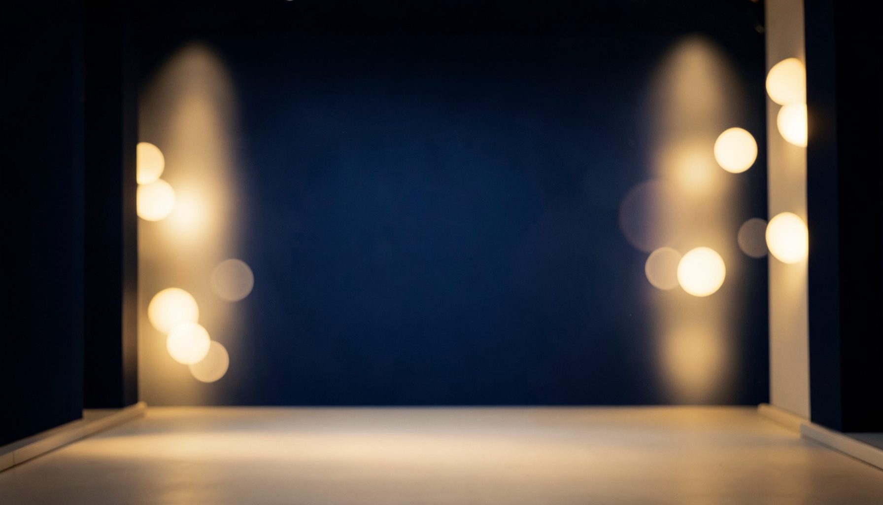 A warm, softly lit entertainment studio backdrop with deep navy and cream tones, featuring abstract bokeh lights and a cinematic atmosphere.