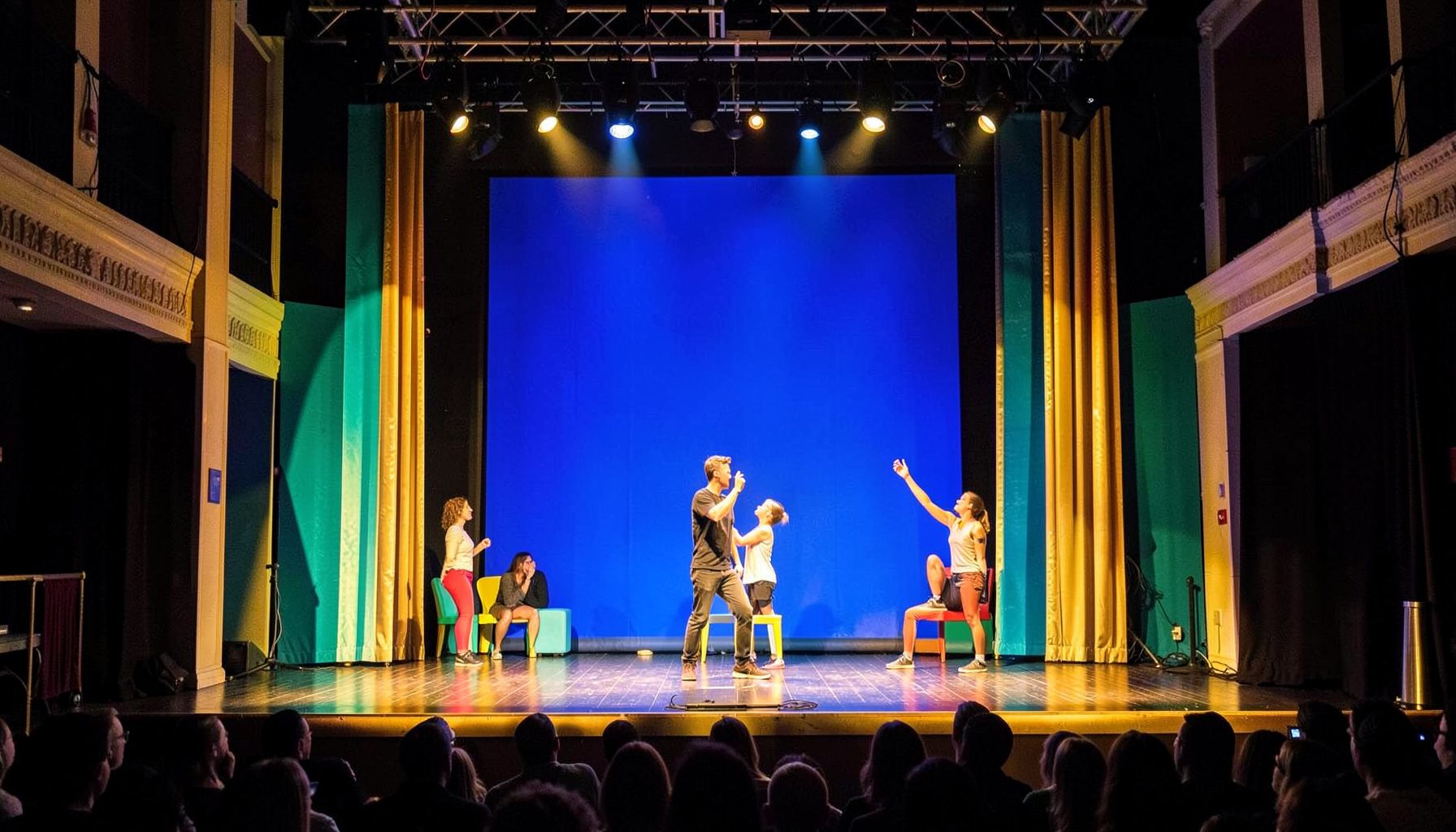 Brightly lit comedy stage with colorful set pieces, warm gold and teal tones, energetic performance atmosphere