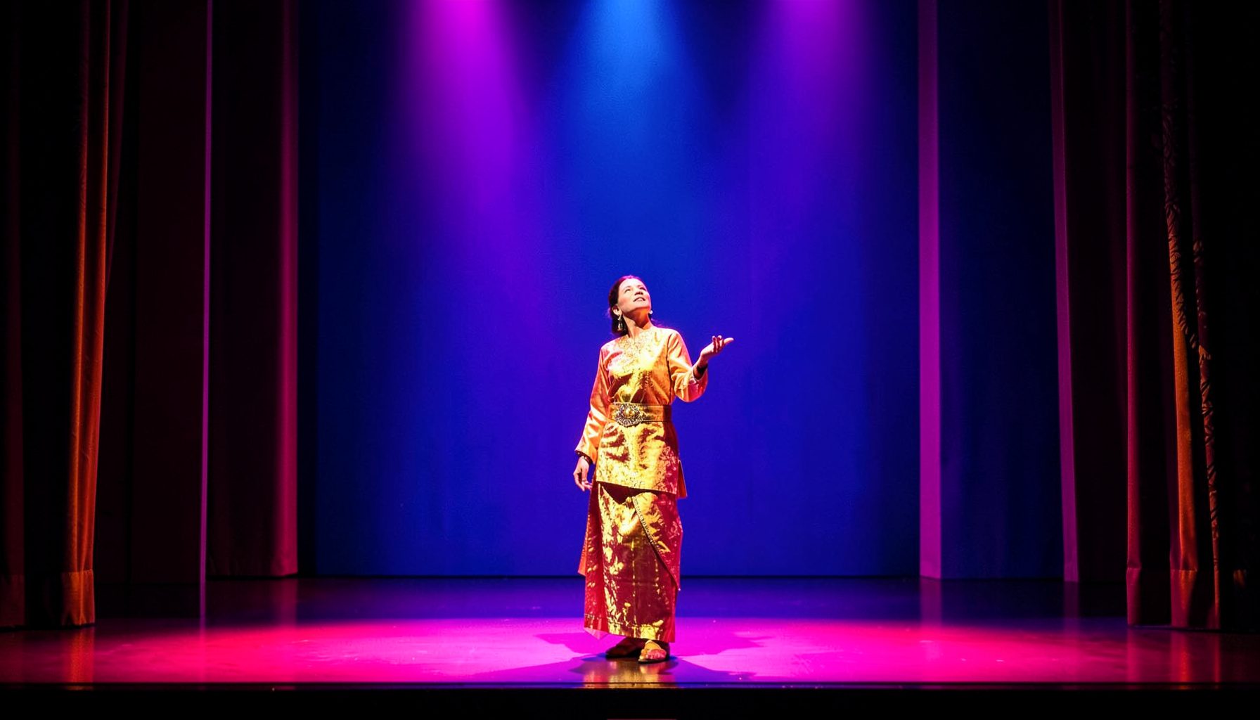 Vibrant stage performance with colorful theatrical lighting in magenta and gold, a single performer in traditional attire mid-gesture