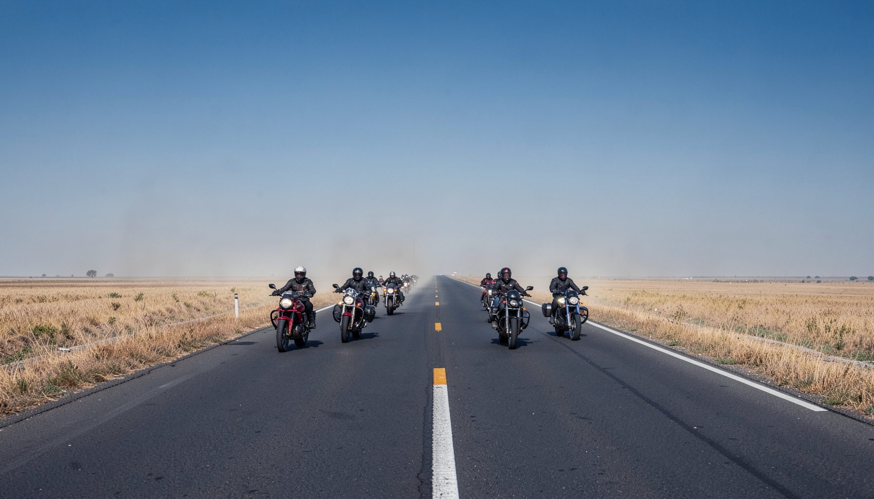 A group of motorcyclists on an open highway with dry grassland stretching to the horizon under a hazy sky. Dust rises faintly behind the bikes.